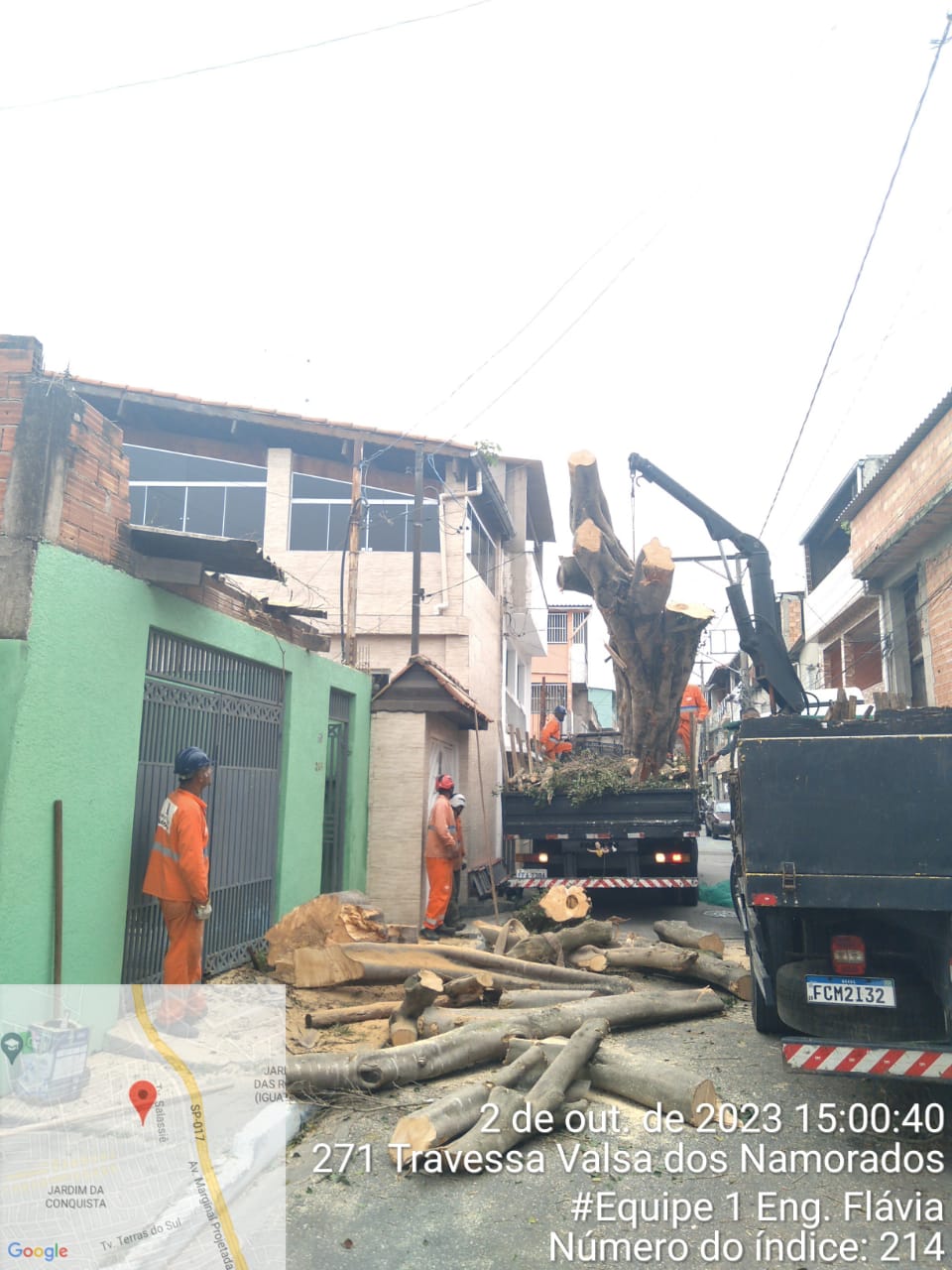 Funcionários da Prefeitura, com macacão laranja, realizaram o corte da árvore e estão ao lado dos galhos recortados, na rua. 